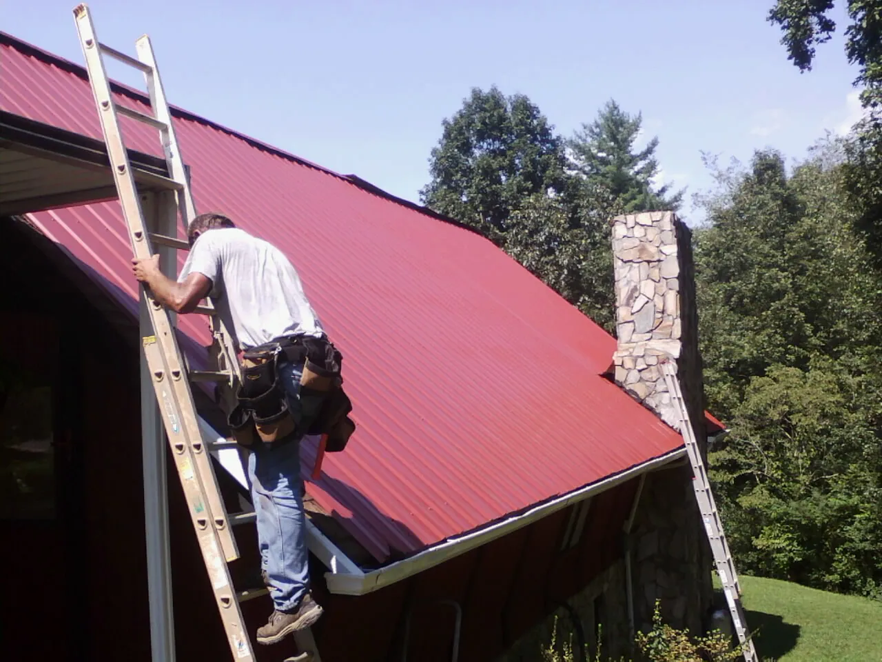 Roofer on a ladder beside a red metal roof and stone chimney — Soffit & Fascia Repair by Roofing Brothers in Sharon Hill